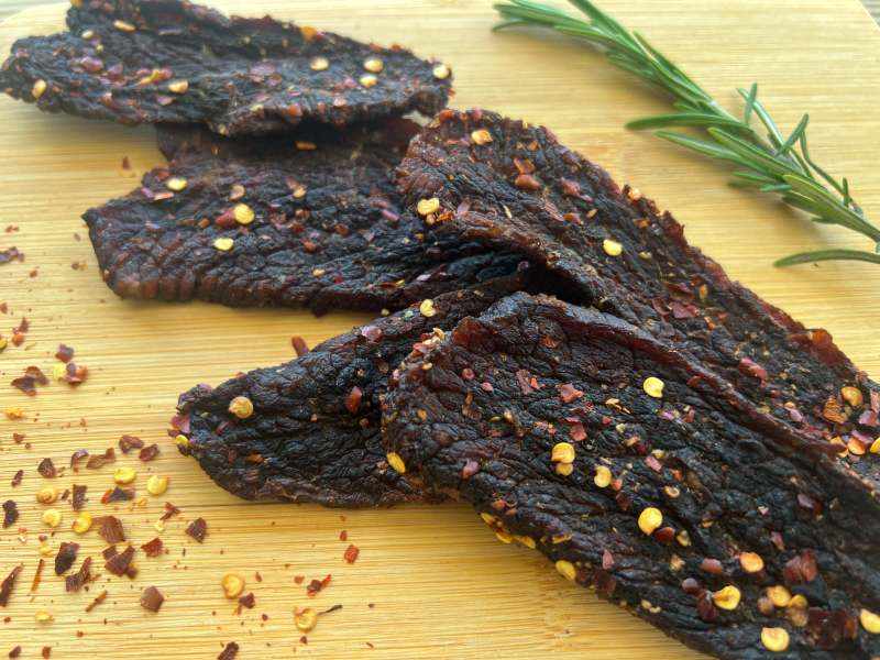 Pepper-speckled jerky pieces on a board beside rosemary