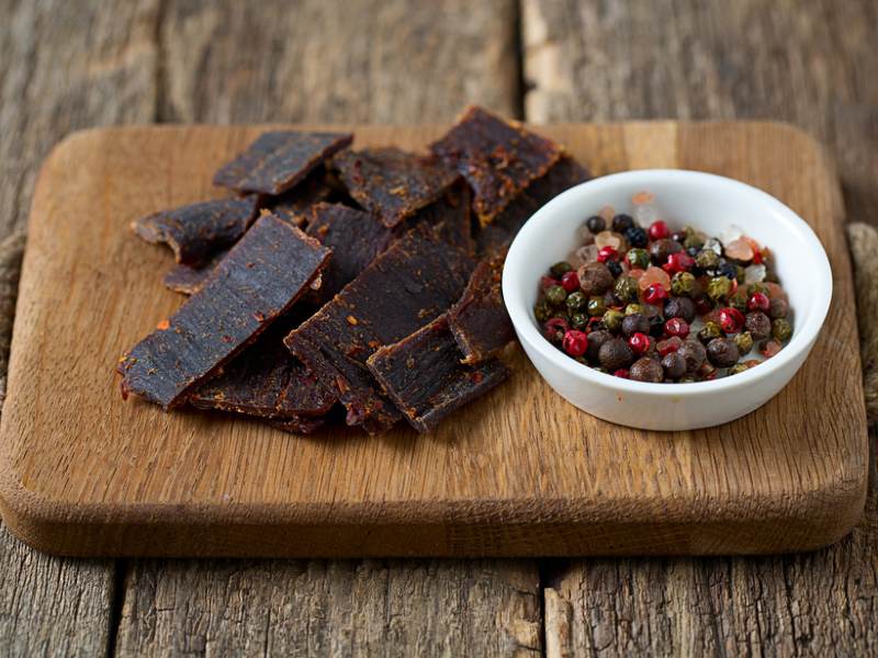 Beef jerky on a board beside a bowl of mixed peppercorns