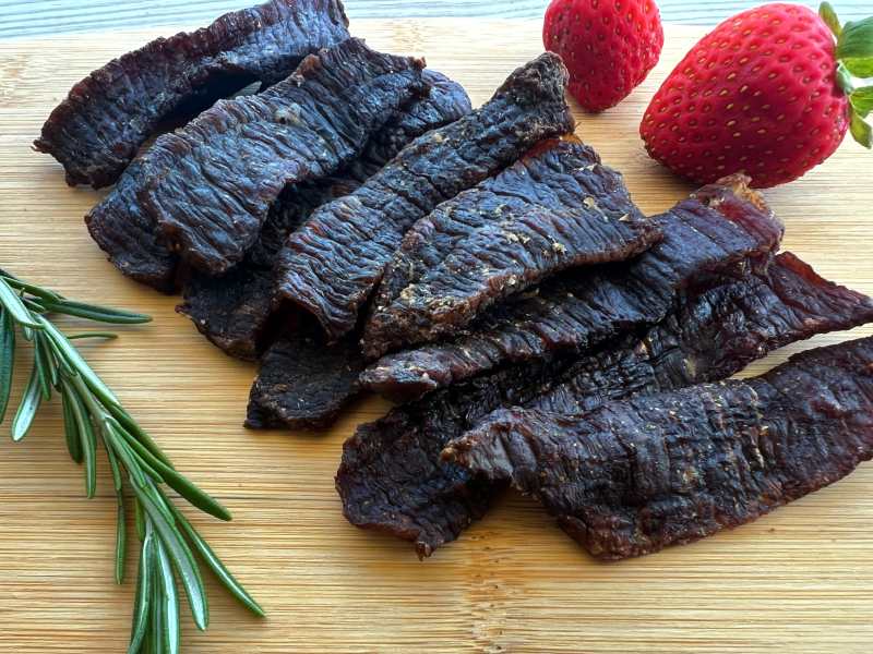 Dark beef jerky strips on a board beside strawberries and rosemary