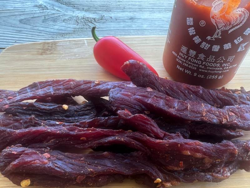 Chili garlic jerky strips beside a red pepper and Sriracha bottle