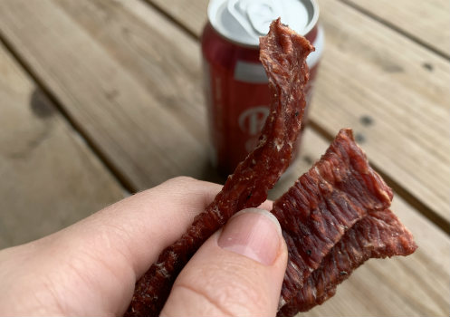 A hand holding a torn strip of jerky beside a Dr Pepper can