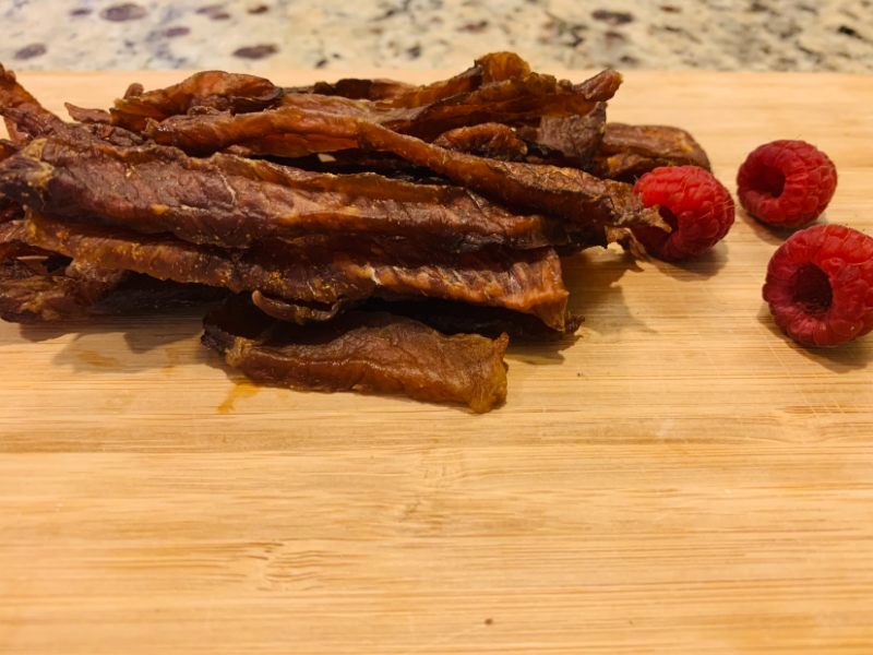 Pork jerky strips piled on a board beside raspberries