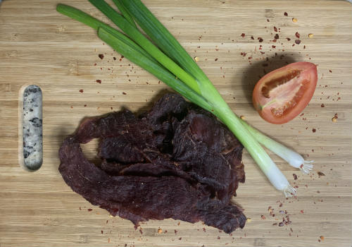 Spicy jerky piled on a board beside scallions and a tomato