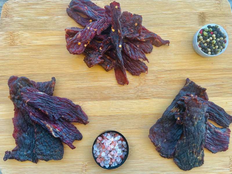 Three piles of jerky beside small bowls of salt and pepper