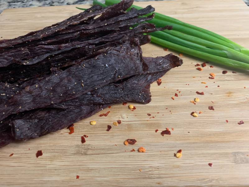 Dark homemade beef jerky strips beside scallions and pepper flakes