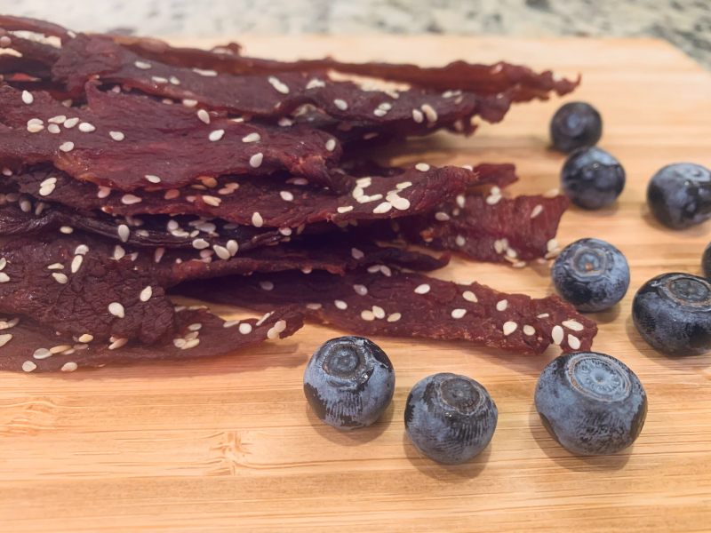 Sesame-coated beef jerky strips beside blueberries