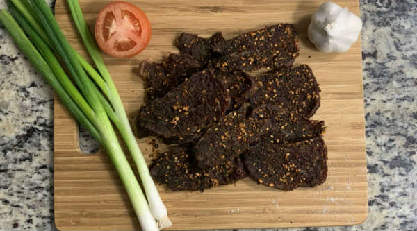 Finished spice-crusted jerky beside scallions, tomato, and garlic
