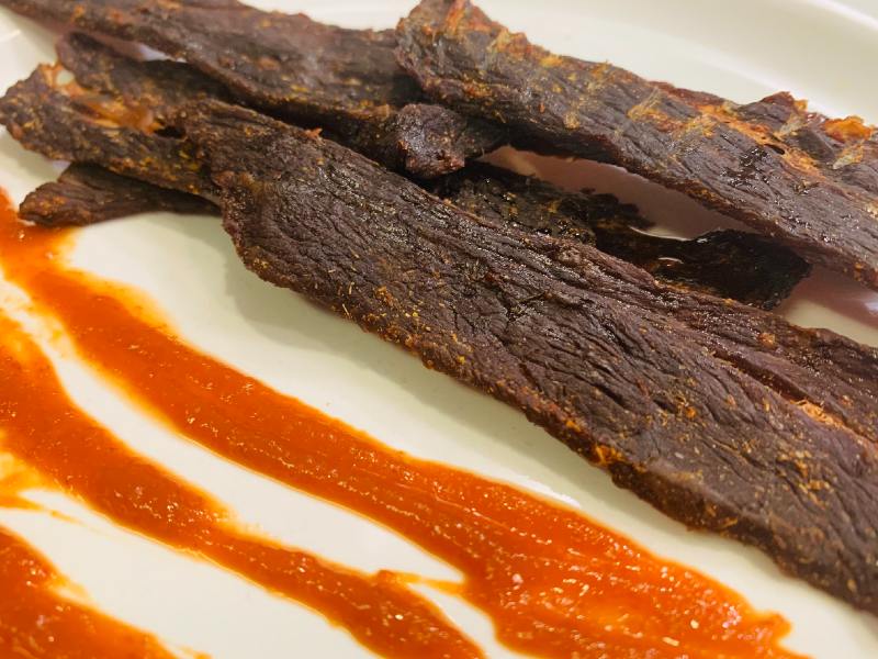 Dark beef jerky on a plate with a streak of red sauce
