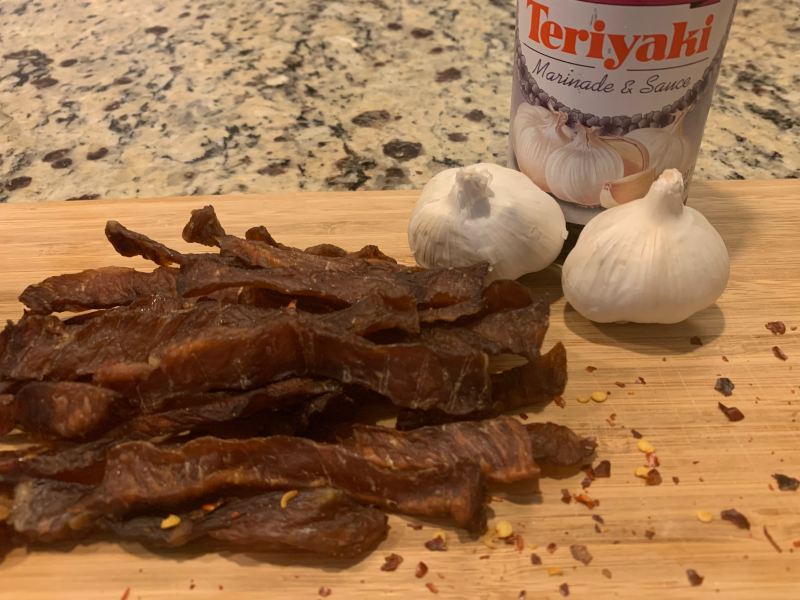 Pork jerky beside garlic bulbs and a bottle of teriyaki marinade