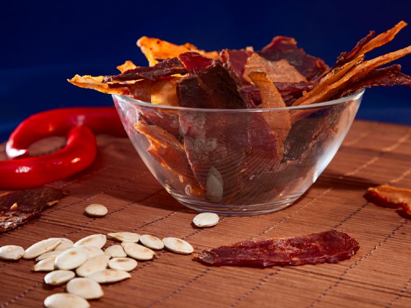 Strips of dried food in a glass bowl beside peppers and seeds