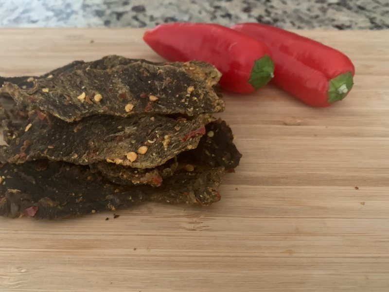 Dark beef jerky coated in herbs and spices beside red peppers